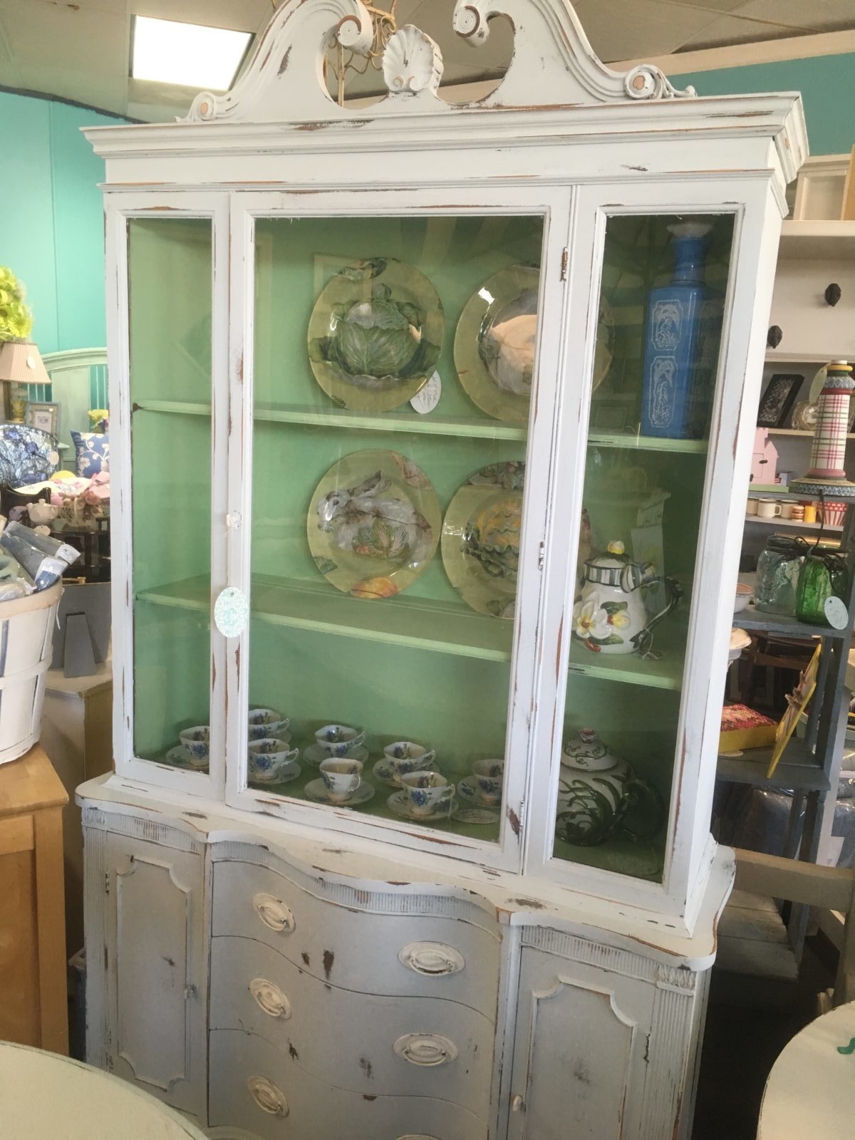 Chalk Paint China Cabinet • Antique china cabinet in a faint blue chalk paint with a pleasing green interior to showcase your china or favorite ceramics. Features an ornate scroll top, plate grooves and storage underneath for silverware & napkins.